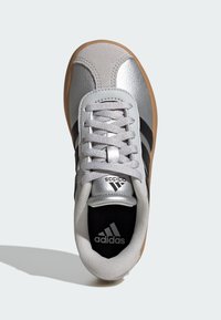 Srebrne atletske tenisice s crnim prugama, teksturiranom gornjom površinom, okruglim prstom i gumiranim potplatima. Imaju vezanje sprijeda i brendiranu unutrašnjost.