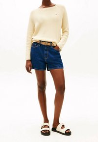 Pull en maille crème, short en denim bleu avec poches avant, ceinture camel avec boucle métallique, et sandales blanches avec semelles noires et accents dorés.