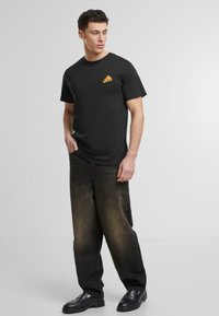 Svart t-skjorte med et lite pizzamotiv på venstre bryst, kombinert med mørke, løstsittende jeans og svarte sko.