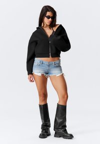 Svart kortad zip-up hoodie, denimshorts med fransade kanter och svarta läderstövlar i knähöjd. Modellen bär mörka solglasögon.