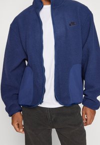 Marinblå fleecejacka med texturerad tyg, dragkedja framtill och två sidofickor. Har en enhetlig blå kant och en liten svart logotyp.