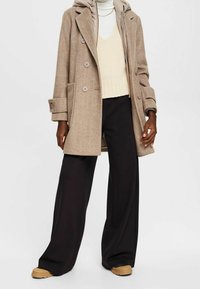 Beżowy płaszcz w jodełkę z dużymi guzikami i kapturem, noszony na kremowym swetrze, w zestawie z czarnymi szerokimi spodniami i brązowymi butami.