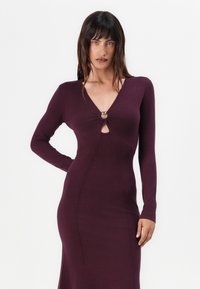 Langärmliges Kleid mit tiefem V-Ausschnitt aus dunkelviolettem Strickstoff, das ein Knotendetail mit einem kreisförmigen Akzent auf der Brust und eine taillierte Silhouette aufweist.