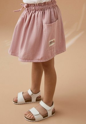 Enfant portant une jupe rose froncée à la taille avec poche appliquée et sandales blanches à velcro, debout sur un sol et un fond beiges.