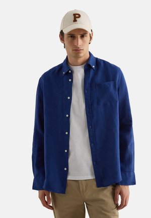 Giovane uomo con un berretto beige, una camicia azzurra con bottoni marrone "P," sopra una maglietta bianca e pantaloni beige, in piedi davanti a uno sfondo uniforme.