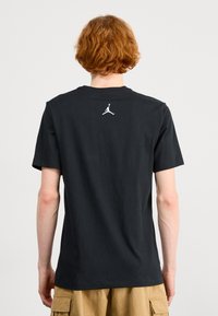 Černé bavlněné tričko s krátkým rukávem, s bílým logem Jumpman na horní části zad. Jednoduchý design s kulatým výstřihem.