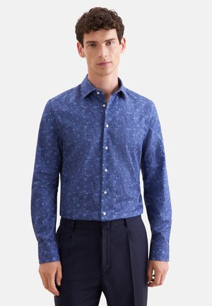 Man draagt een strak blauw bloemenpatroon overhemd met lange mouwen en donkere broek, staand tegen een effen achtergrond.