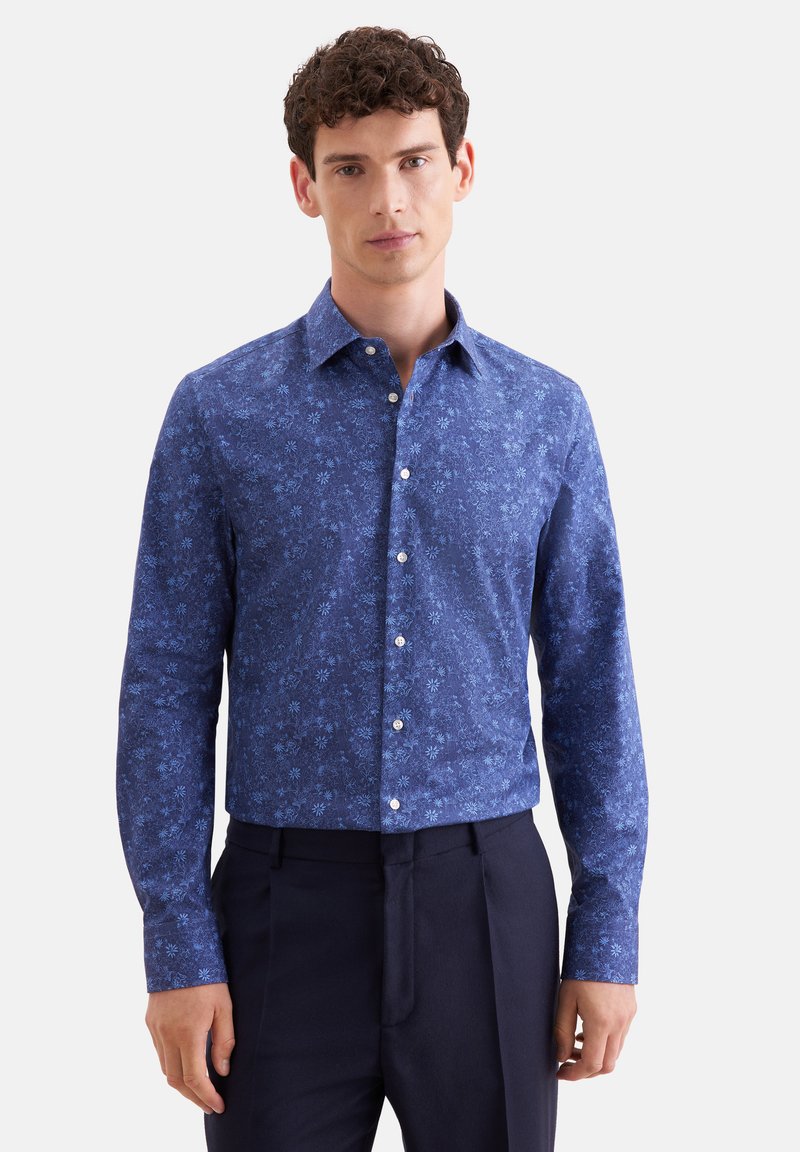 Homme portant une chemise bleue ajustée à fleurs à manches longues boutonnée et un pantalon foncé, debout devant un fond uni.