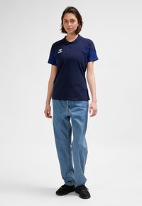 Mörkblå pikétröja med korta ärmar, utrustad med krage och logotyp, kombinerad med ljusblå högmidjade jeans och svarta sneakers.