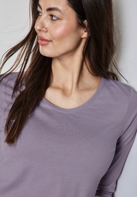 Top lungo a maniche con scollo tondo di colore lilla, texture liscia e design aderente, che si drappeggia dolcemente sulle spalle.