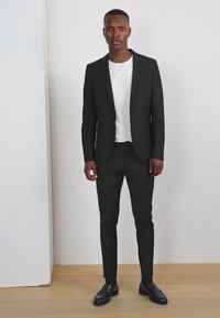 Completo nero su misura con silhouette aderente, abbinato a una t-shirt bianca a girocollo. L'outfit presenta un tessuto morbido e scarpe nere eleganti.