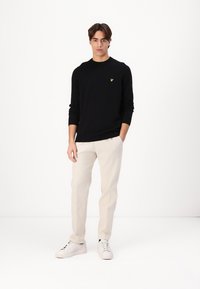 Zwarte gebreide trui met lange mouwen en een ronde hals, voorzien van een klein geel logo. Gepaard met lichtbeige broek en witte sneakers.