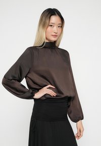 Young woman with long ombre hair wearing a dark brown satin blouse with puffed sleeves and a high-waisted black skirt, posing with hand on hip.