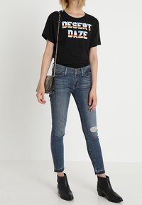 Camiseta negra de manga corta con el texto multicolor "DESERT DAZE", combinada con jeans skinny de lavado medio en azul y botas negras hasta el tobillo. Bolso de mano con estampado de serpiente.