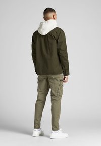 Olijfgroene jas met een witte capuchon, gecombineerd met licht olijfgroene cargobroeken en witte sneakers. De stof lijkt stevig en heeft een ontspannen pasvorm.