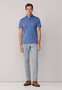 Polo bleu à manches courtes avec col, associé à un pantalon gris clair et des chaussures marron. Le mannequin se tient devant un fond neutre.