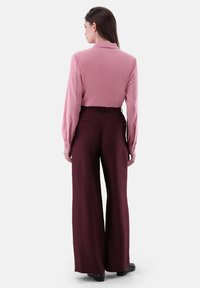 Langärmlige pinke Bluse mit Kragen, kombiniert mit hochgeschnittenen, lässigen burgunderfarbenen Hosen. Glatte Textur und minimale Details.