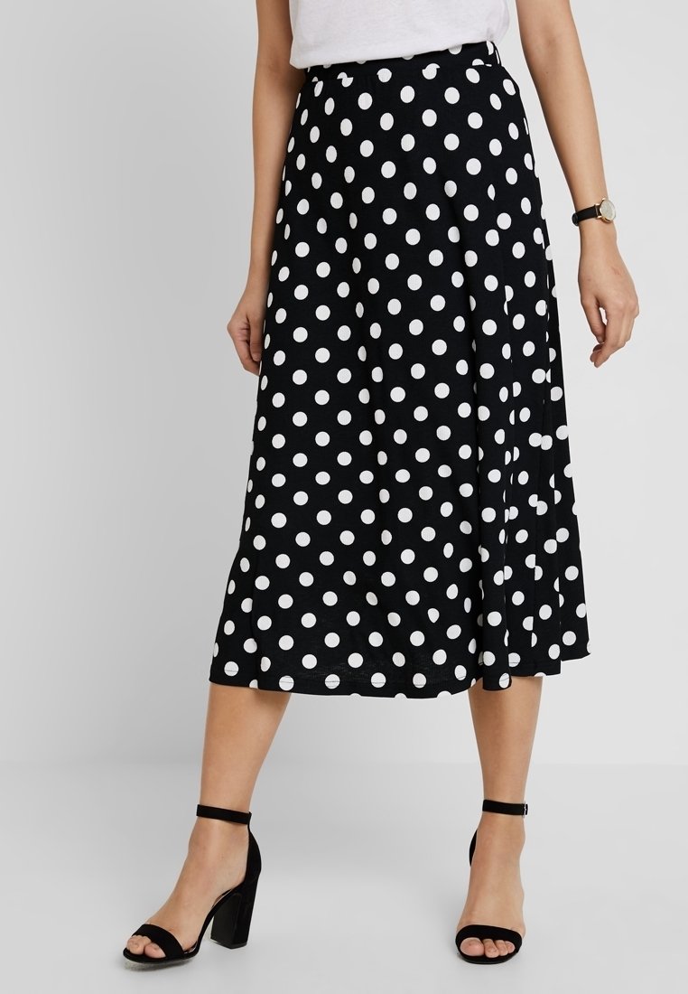 Femme portant une jupe midi noire à pois blancs, un haut blanc, des talons noirs à bride à la cheville et une montre-bracelet au poignet gauche.