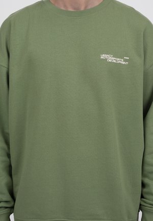 Man wearing olive green sweatshirt with white text "Legacy Motorsports Development 2022" on left chest, shown from neck to waist.