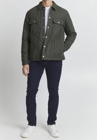 Přesnívaná olivově zelená bunda se cvoky, dvěma náprsními kapsami a límečkem, v kombinaci s tmavými džíny a bílými teniskami.