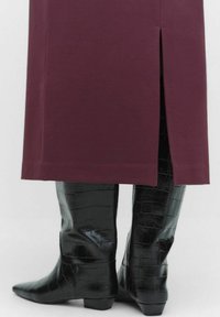 Jupe bordeaux avec une fente à l'arrière, associée à des bottes noires brillantes montantes en cuir croco à bout pointu.