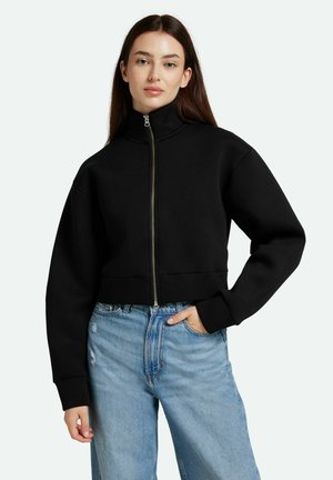 Sudadera negra corta con alto cuello y cremallera frontal, hecha de material suave. Hombros anchos y caídos y puños acanalados. Jeans de color azul claro.