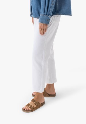 Osoba ubrana w białe krótkie spodnie, brązowe sandały z podwójną klamrą oraz niebieską dżinsową kurtkę, stojąca z rękami swobodnie opuszczonymi wzdłuż ciała.