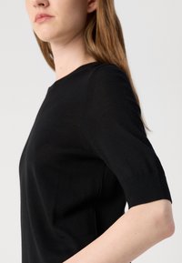 Svart strikket topp med rund halslinning og korte ermer med ribbestrikket mansjett. Glatt tekstur og tettsittende design, som viser subtile sømmer.