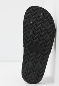 Semelle en caoutchouc noir avec un motif zébré texturé et des grips circulaires. Présente une étiquette logo en relief pour plus de détails.