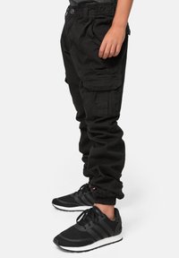 Barn i svarta cargobyxor med elastiska cuffs och svarta sneakers med vita sulor, stående mot en vit bakgrund.