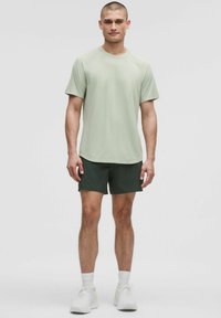 Hellgrünes Sportshirt mit kurzen Ärmeln und strukturierter Oberfläche, kombiniert mit dunkelgrünen Shorts und weißen Sportschuhen.