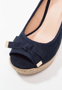 Chaussure compensée bleu marine à bout ouvert avec un nœud en tissu aux extrémités dorées et une semelle en corde de jute sur fond blanc.