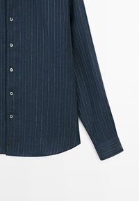 Marineblå langærmet skjorte med knapper, tynde hvide lodrette nålestriber og knaplukket manchet.