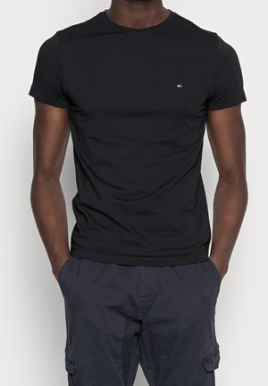 Un homme portant un t-shirt noir ajusté à manches courtes avec un petit logo rouge, blanc et bleu sur la poitrine, et un pantalon cargo sombre avec les mains dans les poches.