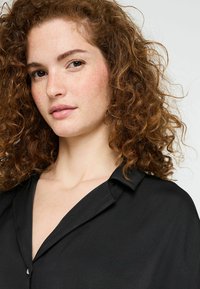 Chemise noire avec col, fermeture à boutons et tissu lisse et doux. Cheveux bouclés et taches de rousseur visibles, avec un fond neutre.