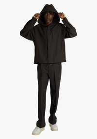 Uomo con giacca nera con cappuccio e pantaloni abbinati, entrambi larghi, che sistema il cappuccio, in piedi su sfondo bianco, indossa scarpe da ginnastica bianche con suola beige.