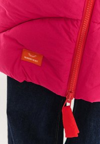 Růžová prošívaná bunda na zip s červeným jezdcem. Má oranžovou visačku s nápisem „ZAHRNUJEŠ TO NEJLEPŠÍ“ přišitou na ní.