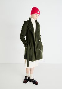 Abrigo de lana verde mezcla con cuello entallado, bolsillos laterales y detalles de cremallera. Llevado sobre un vestido de punto color crema, combinado con un gorro rojo y zapatos negros.