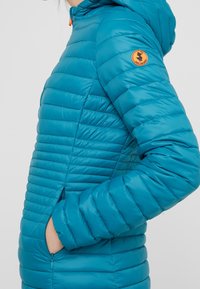 Persona viste una chaqueta de plumas acolchada en color azul verdoso con capucha y una insignia circular naranja en la parte superior del brazo, con la mano en el bolsillo con cremallera.