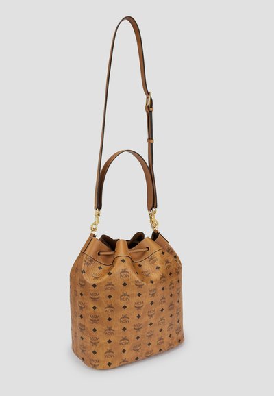 Sac seau en cuir marron avec motif de logo noir, bandoulière longue ajustable et poignée plus courte, posé sur un fond gris clair uni.