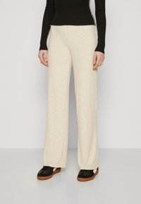 Pantaloni beige a coste, dal taglio ampio e texture liscia, con vita alta, abbinati a scarpe nere con un contrasto bianco.
