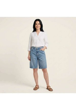 Kvinde med sort hår iført hvid langærmet skjorte, knælange denimshorts og brune sandaler, står med den ene hånd i lommen.