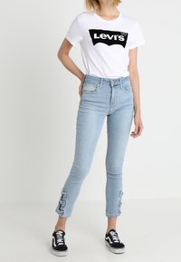 Vit t-shirt med svart Levi's-logotyp, ljusblå skinny jeans med rosett-accenter vid bensluten, tillsammans med svart och vit Vans-sneakers.