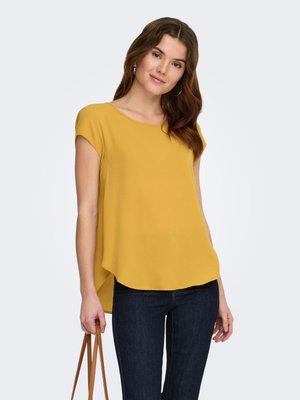 Woman wearing mustard yellow short-sleeve blouse and dark jeans, holding brown handbag straps in her left hand, against plain light background.