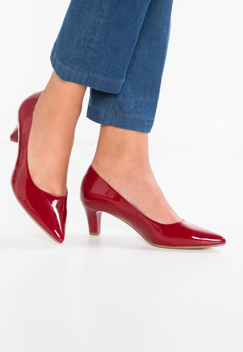 Femme portant des escarpins rouges brillants à talons bas et bouts pointus, avec un jean bleu raccourci, debout sur un sol blanc.