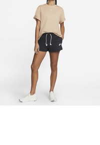 Beige bomullströja med rullade ärmar, tillsammans med svarta shorts med dragsko och vitt logotyp, samt vita strukturerade sneakers.