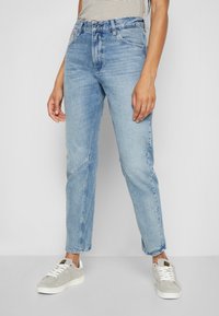 Jeans de talle alto en azul claro con un ajuste relajado, textura desgastada y dobladillos cortados, combinados con zapatillas de ante grises y una blusa a rayas.