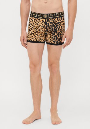Mann in Leoparden-Print Boxershorts mit schwarzem und goldenem Bund, der ein geometrisches und medaillonartiges Design aufweist, steht barfuß vor weißem Hintergrund.