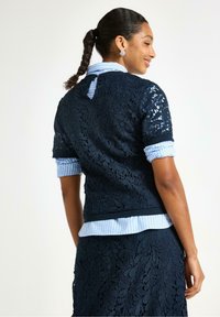 Marinblå blondetop med blomsterprint, korte ærmer og detalje med nøglehul i ryggen, lagdelt over en lyseblå stribet skjorte.