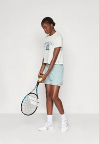 Naine valges Monaco T-särgis ja heledates sinistes lühikeses seelikus, hoides tennisereketit ja -palli, seismas lihtsa valge tausta ees.